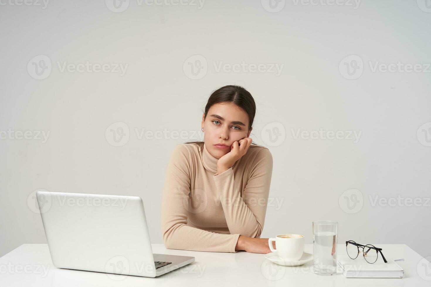 intérieur photo de Jeune épuisé aux yeux bleus brunette femelle en portant sa tête avec élevé main et à la recherche fatigué à caméra, ayant difficile journée à travail, isolé plus de blanc Contexte