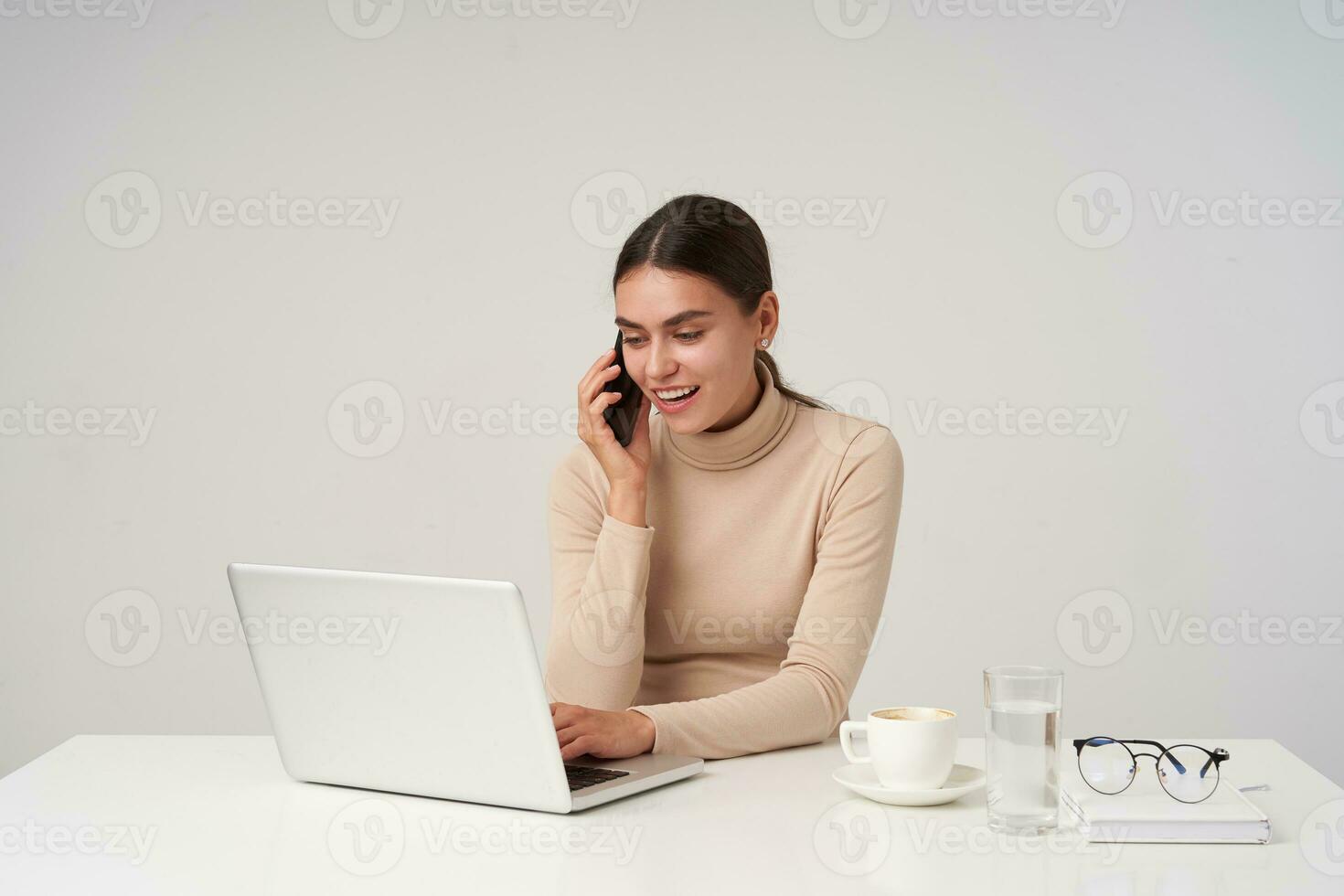 coup de Jeune jolie positif foncé aux cheveux Dame avec Naturel maquillage souriant gaiement tandis que fabrication appel et à la recherche à écran tandis que dactylographie texte sur clavier, isolé plus de blanc Contexte photo