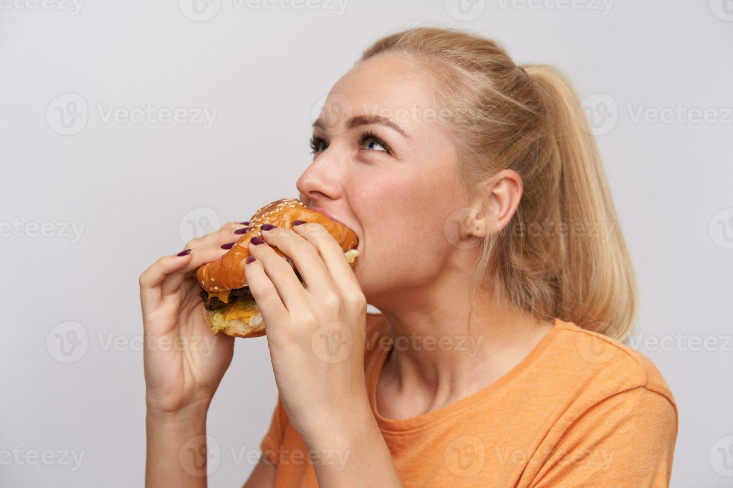 fermer de Jeune magnifique positif blond femme avec décontractée coiffure ravissant Frais savoureux hamburger permanent plus de blanc Contexte dans décontractée T-shirt photo