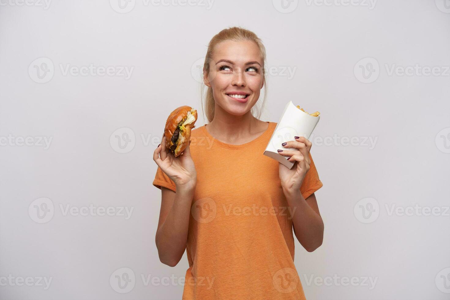 studio coup de Jeune Enchanté blond femelle avec décontractée coiffure en gardant Hamburger et français frites dans sa mains et à la recherche gaiement de côté, mordant lèvre inférieure et avant-goût délicieux dîner photo