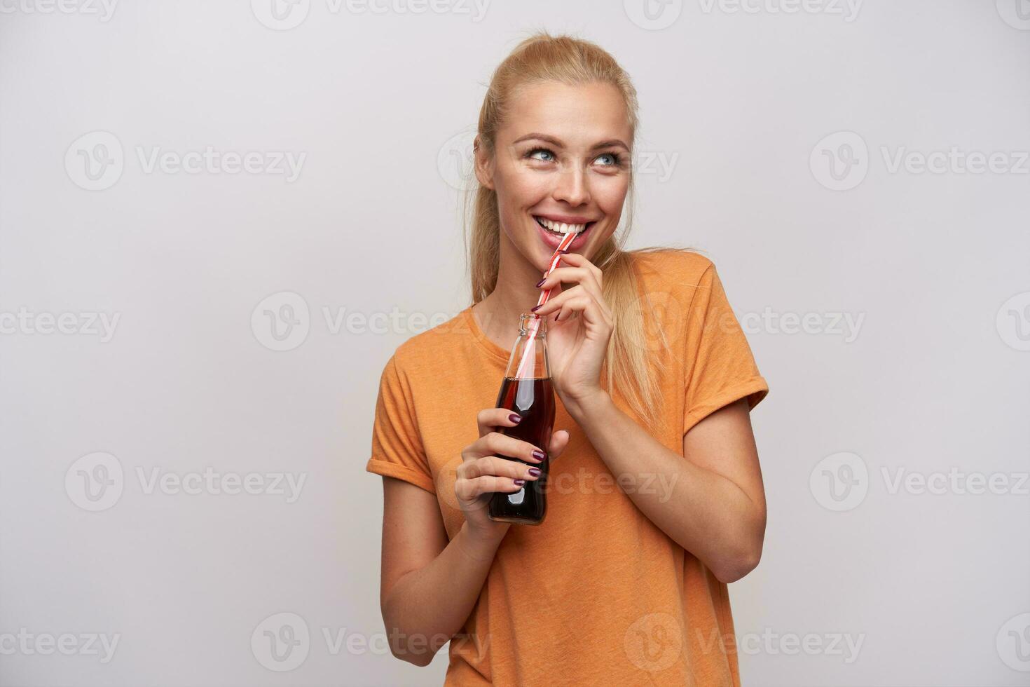 intérieur photo de Jeune joyeux blond femelle avec queue de cheval coiffure en portant verre bouteille de un soda dans élevé mains et à la recherche gaiement de côté, isolé plus de blanc Contexte