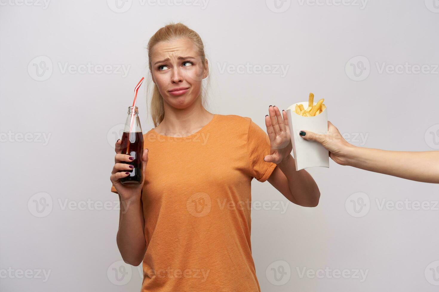 mécontent Jeune blond femelle avec décontractée coiffure à la recherche de côté avec moue et renfrogné sa visage avec élevé palmier, en buvant un soda et refusant à manger français frites, isolé plus de blanc Contexte photo