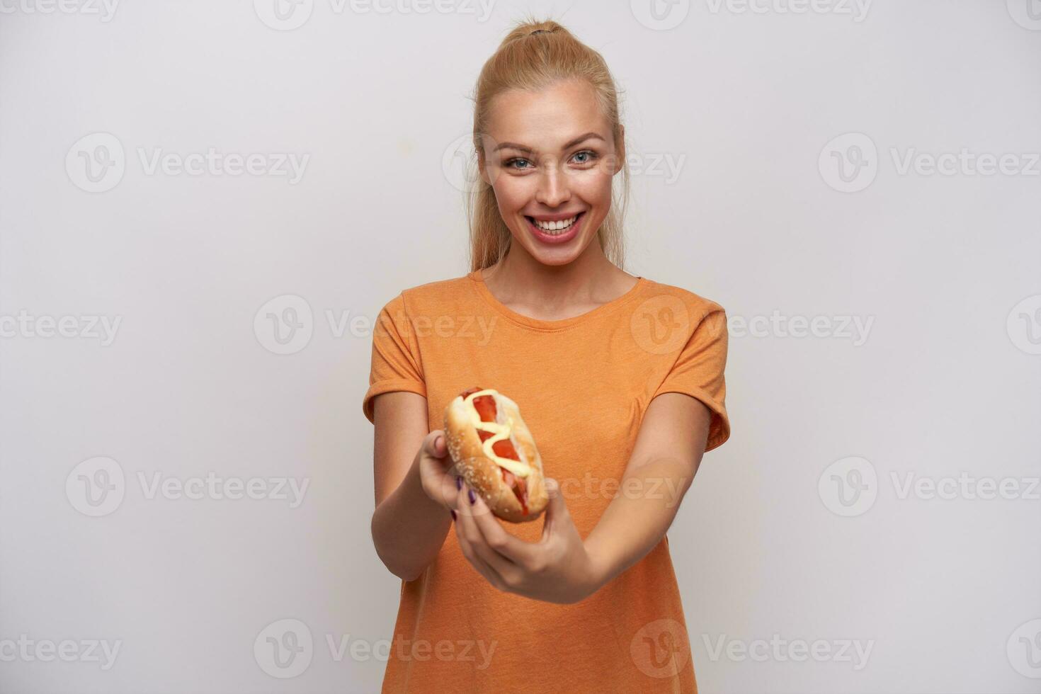 content attrayant Jeune longue aux cheveux blond femme dans Orange T-shirt en portant chaud chien et montrant il à caméra, souriant Cordialement tandis que posant contre blanc Contexte photo