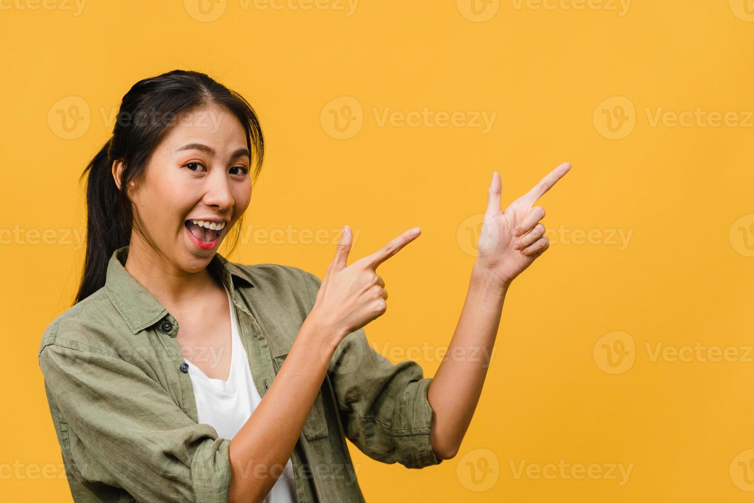 portrait d'une jeune femme asiatique souriante avec une expression joyeuse, montre quelque chose d'étonnant dans un espace vide dans un tissu décontracté et regardant la caméra isolée sur fond jaune. concept d'expression faciale. photo