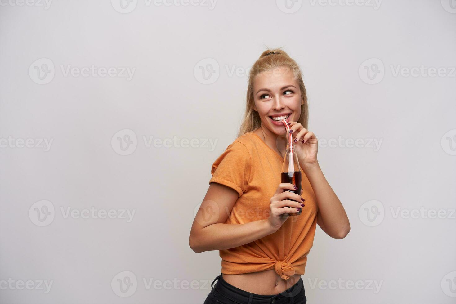 studio photo de de bonne humeur Jeune attrayant femme dans Orange T-shirt en buvant un soda avec paille tandis que à la recherche de côté avec charmant sourire, posant contre blanc Contexte