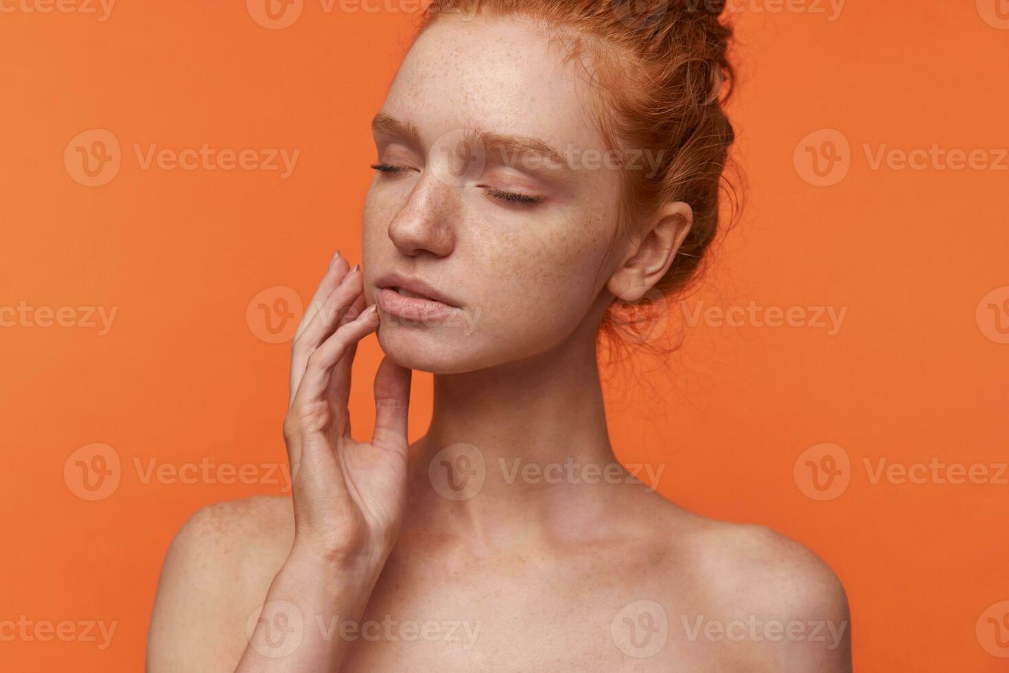 studio portrait de Jeune attrayant tête de lecture femme portant cheveux dans décontractée coiffure posant plus de Orange Contexte avec fermé yeux, émouvant chèrement sa visage avec les doigts photo