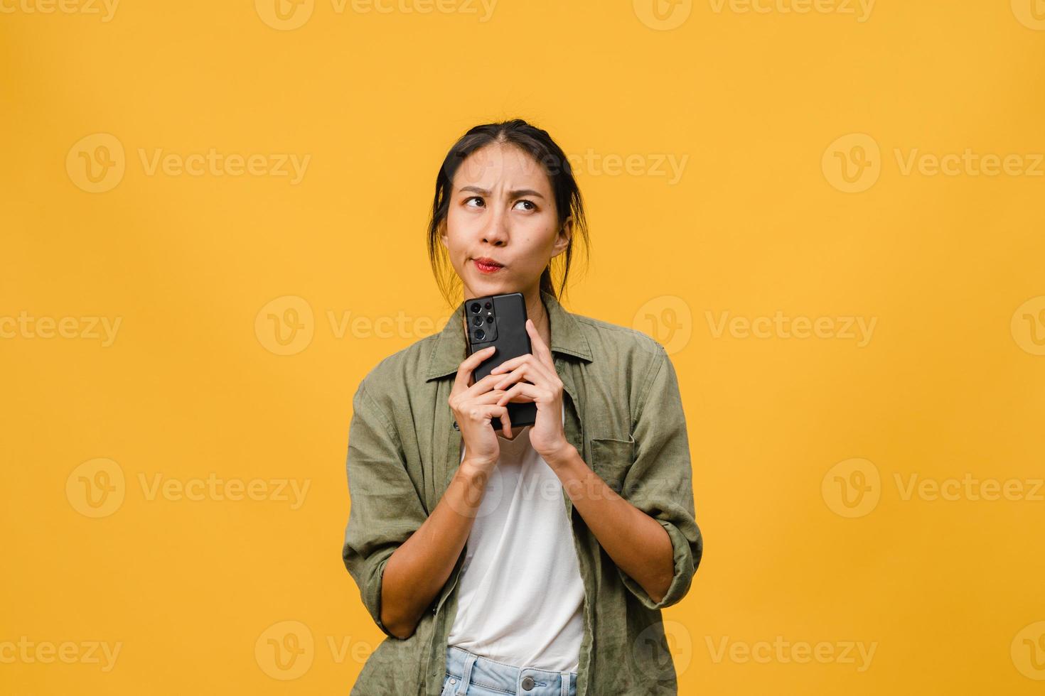 pensant rêver d'une jeune femme asiatique utilisant un téléphone avec une expression positive, vêtue d'un tissu décontracté, se sentant heureuse et se tenant isolée sur fond jaune. heureuse adorable femme heureuse se réjouit du succès. photo