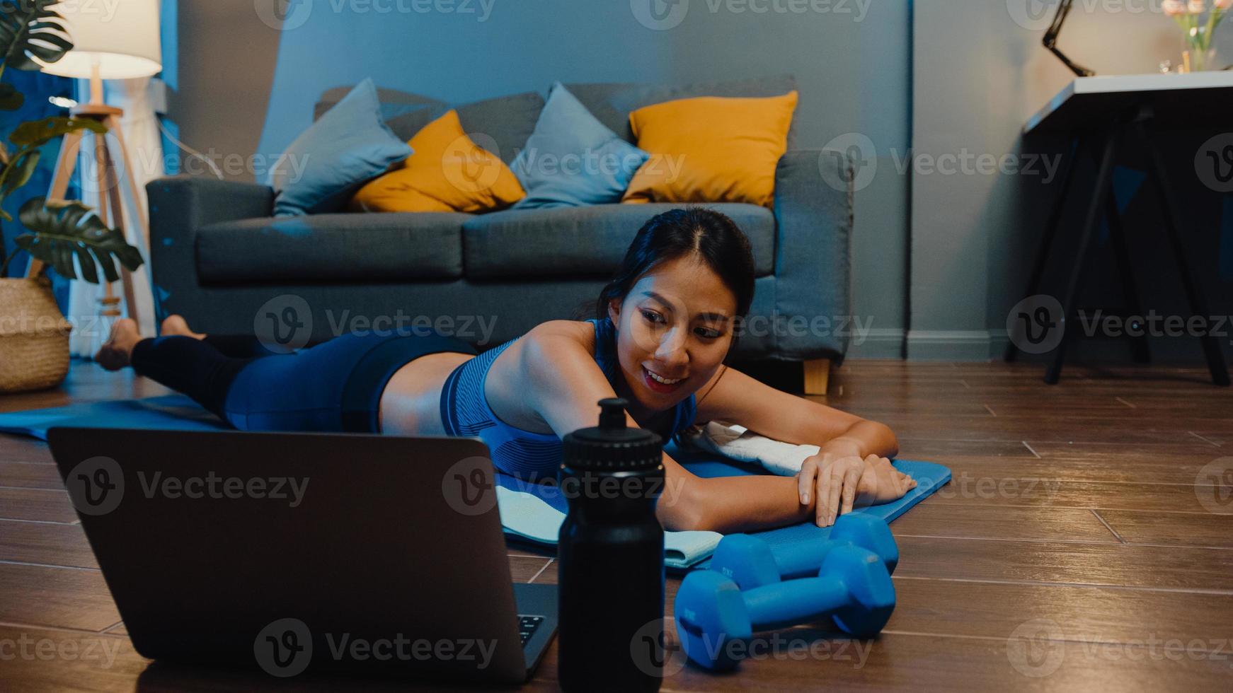 jeune femme asiatique en vêtements de sport faisant de l'exercice et utilisant un ordinateur portable pour regarder un didacticiel vidéo de yoga la nuit à la maison. formation à distance avec entraîneur personnel, distance sociale, concept d'éducation en ligne. photo