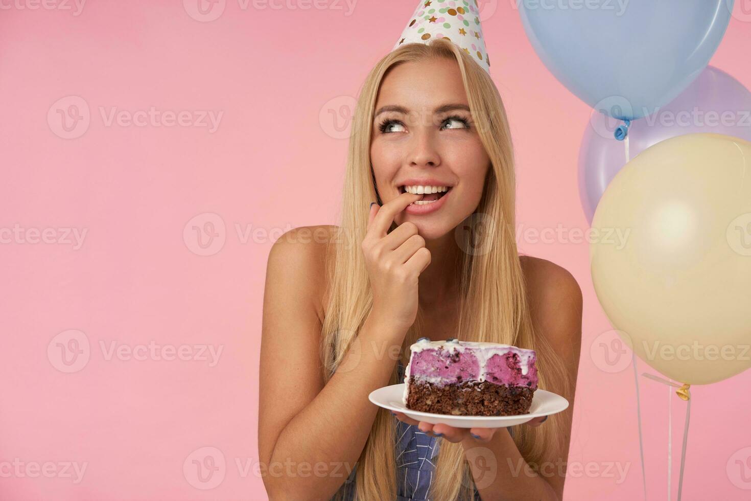 Enchanté jolie femme réjouissance tandis que posant dans multicolore air des ballons, célébrer anniversaire avec délicieux gâteau, à la recherche de côté positivement et rêver à propos avenir, isolé plus de rose Contexte photo