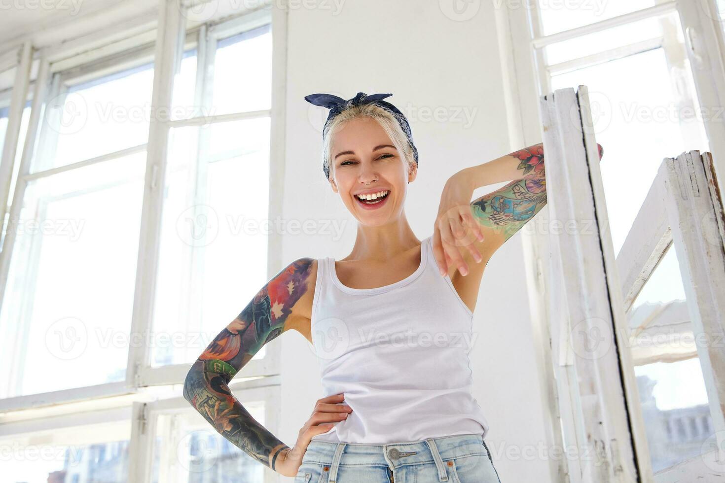 content Jeune magnifique tatoué blond Dame avec décontractée coiffure penché sur en bois fenêtre tandis que à la recherche Heureusement à caméra, permanent plus de brillant studio sur ensoleillé journée photo