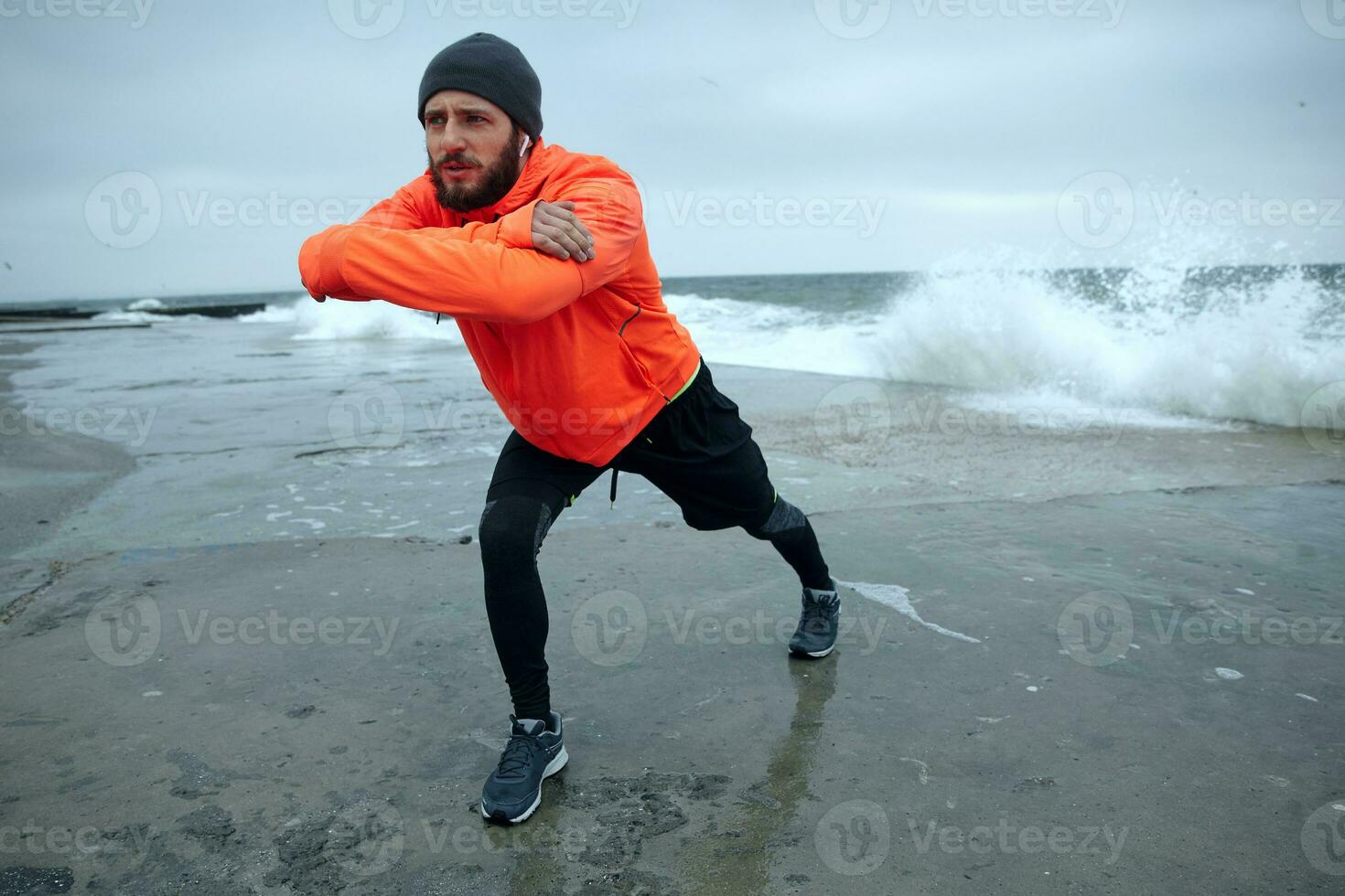 Jeune foncé aux cheveux Beau barbu modèle avec écouteurs en gardant mains dans de face de lui-même et renfrogné les sourcils tandis que à la recherche devant, Faire Matin faire des exercices plus de bord de mer photo