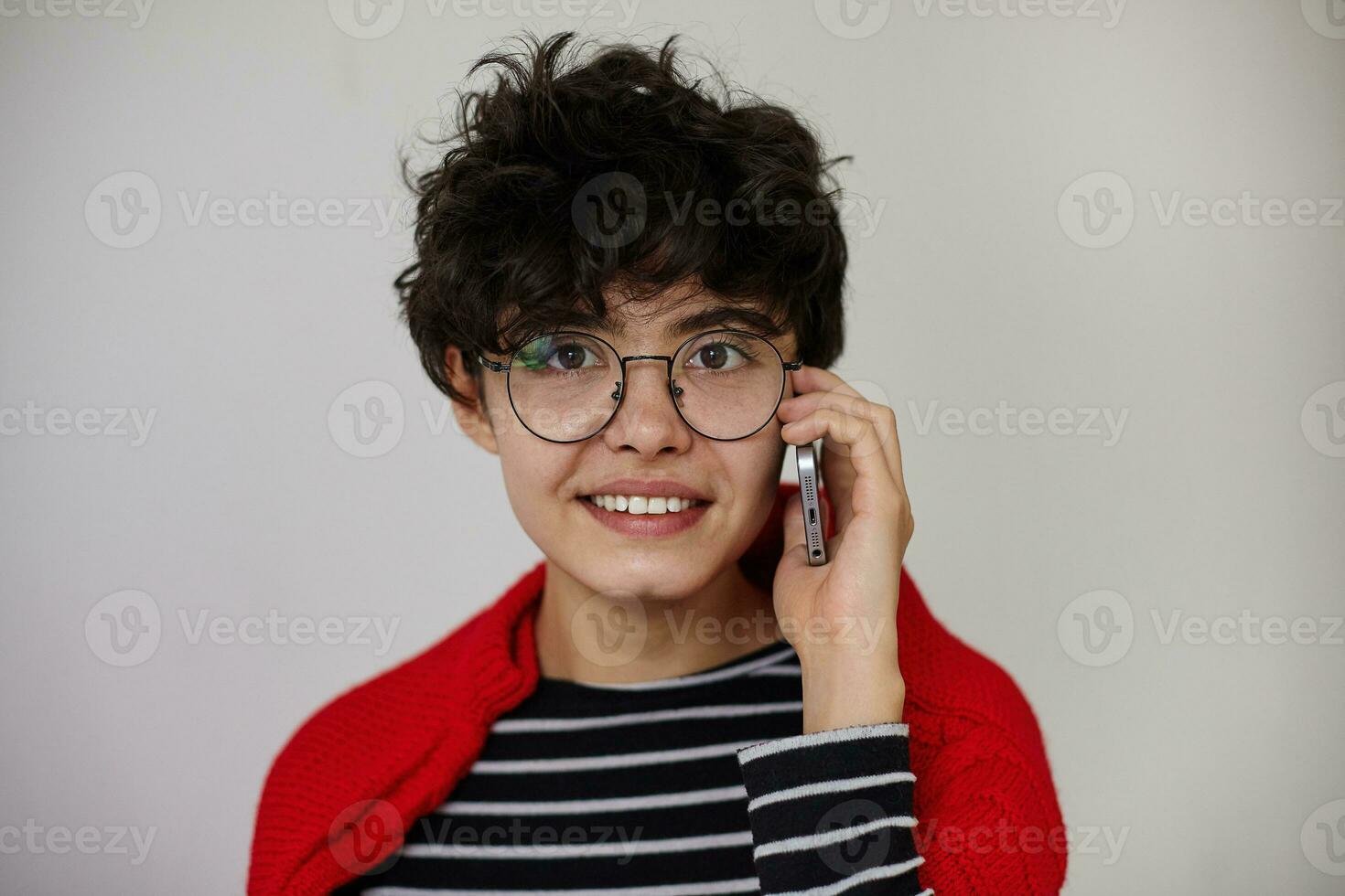 portrait de Jeune aux yeux bruns frisé brunette femelle avec Naturel maquillage fabrication appel avec sa téléphone intelligent et à la recherche positivement à caméra, permanent contre blanc Contexte photo