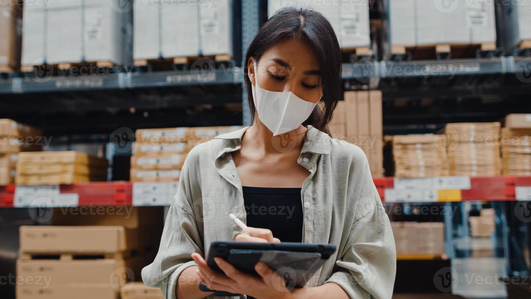 une jeune femme d'affaires asiatique porte un masque facial à la recherche de marchandises à l'aide d'une tablette numérique vérifiant les niveaux d'inventaire dans le centre commercial de détail. distribution, logistique, colis prêts à être expédiés. photo