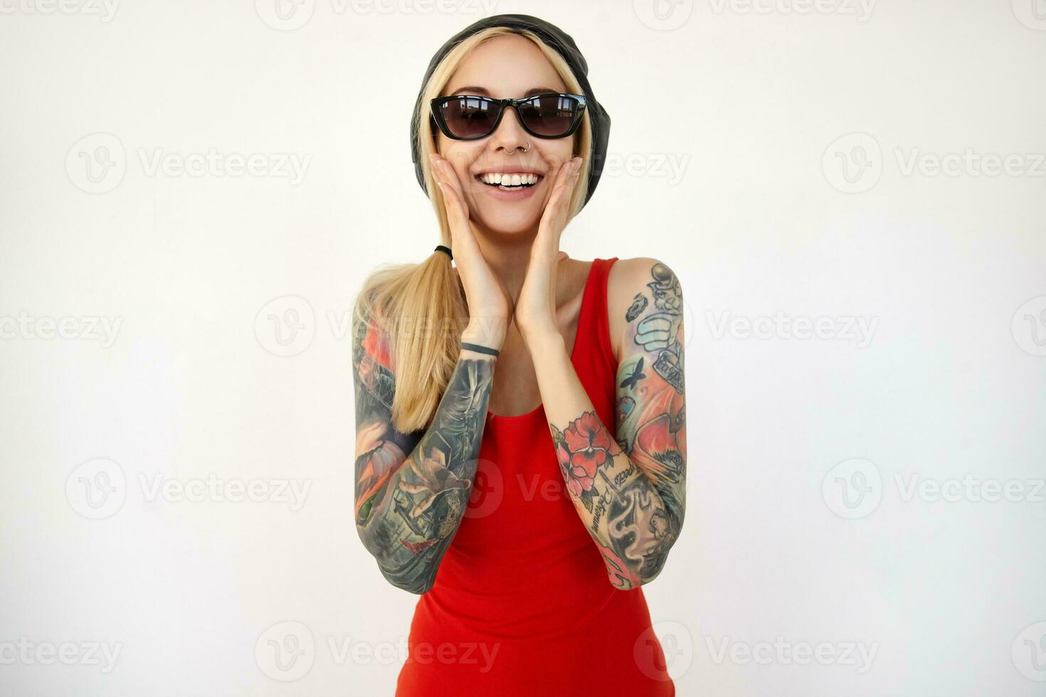 intérieur coup de Jeune attrayant tatoué blond femme en portant sa visage avec élevé paumes et souriant Heureusement à caméra, permanent plus de blanc Contexte photo