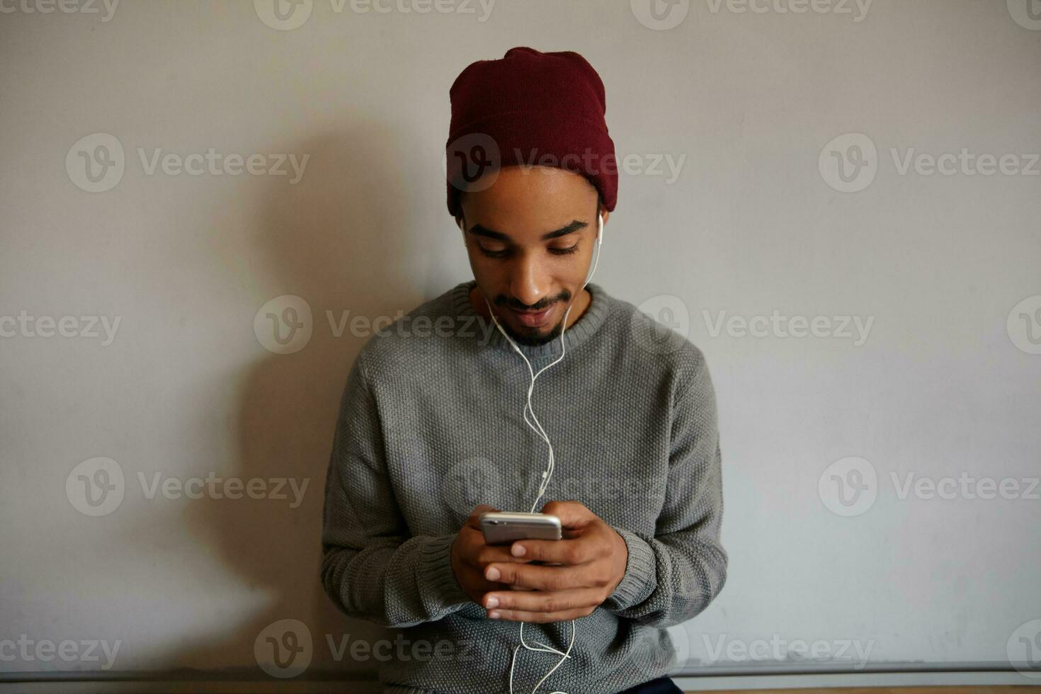 agréable à la recherche jolie barbu gars avec foncé peau habillé dans gris chandail et Bourgogne casquette en gardant mobile téléphone dans élevé mains, bavardage avec copains tandis que séance plus de blanc mur photo