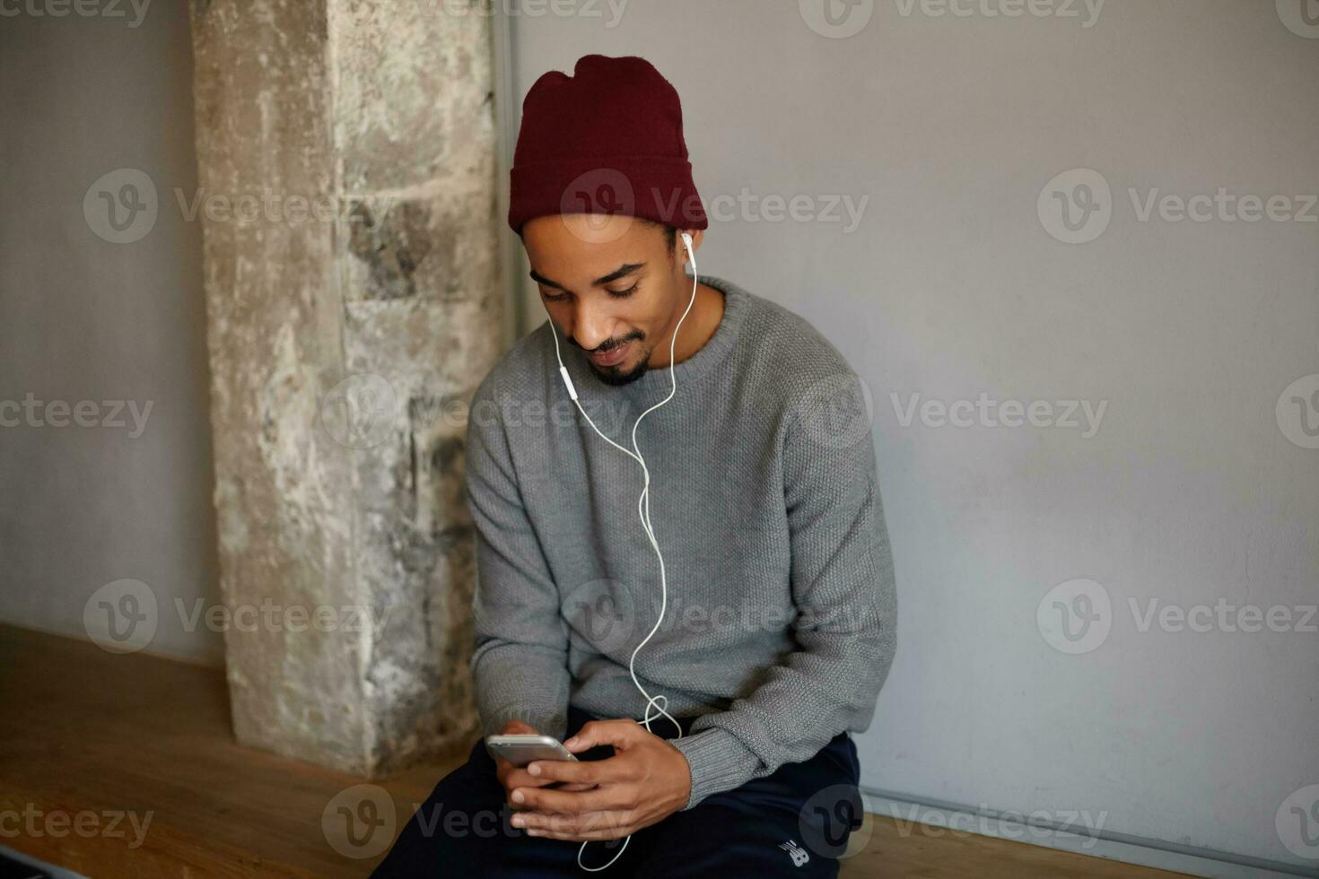 bien à la recherche Jeune barbu foncé écorché gars séance sur longue en bois banc avec écouteurs et mobile téléphone, bavardage avec amis, habillé dans gris chandail et Bourgogne coiffure photo