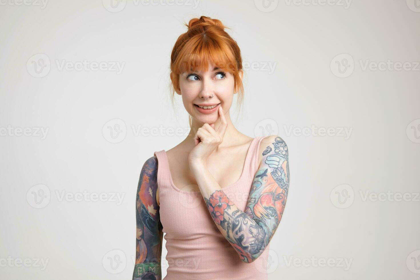 perplexe Jeune jolie roux tatoué Dame avec habillé dans décontractée porter souriant agréablement tandis que à la recherche pensivement de côté, isolé plus de blanc Contexte photo