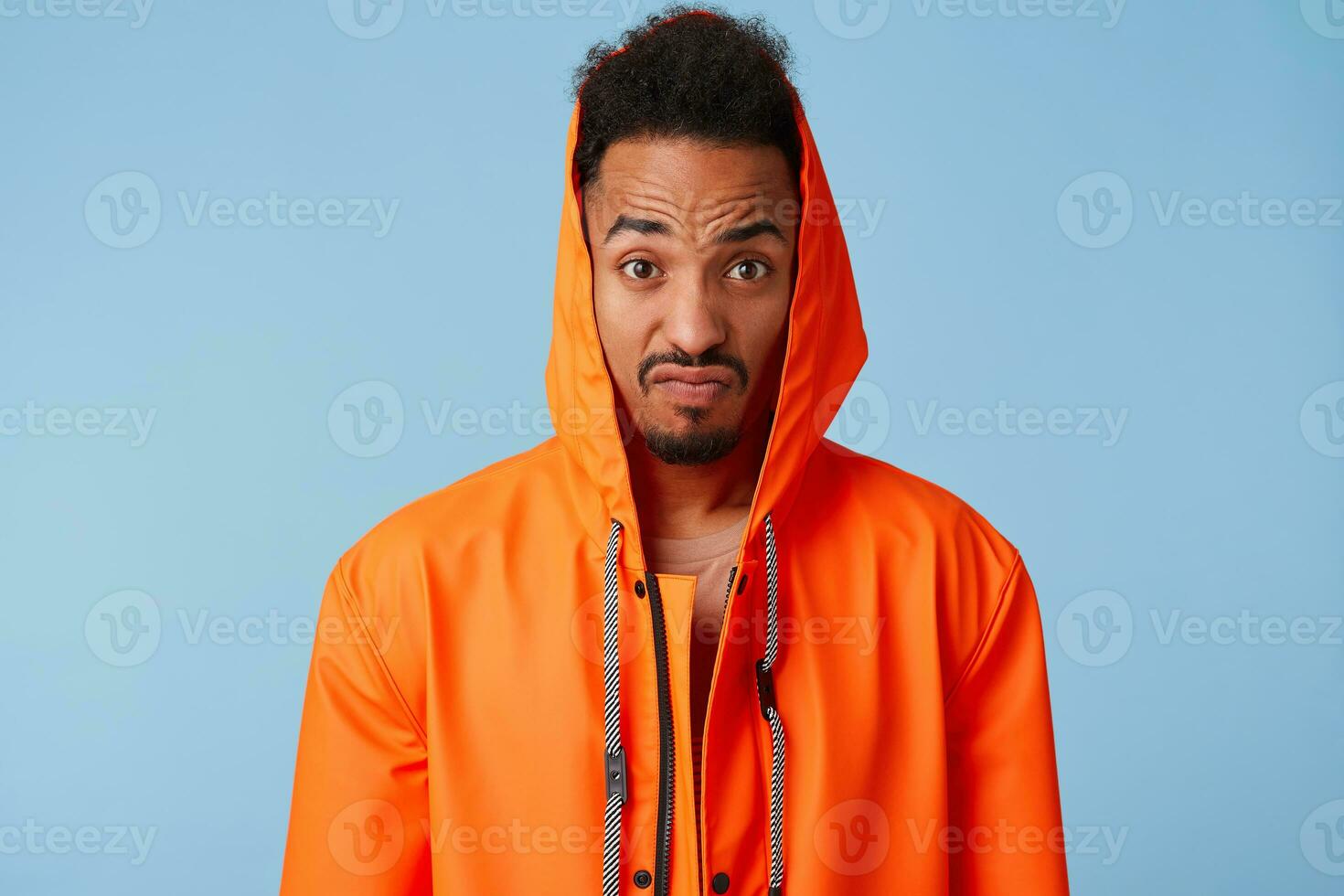 indigné africain américain gars dans Orange pluie manteau se rétrécit le sien les sourcils et serre le sien lèvres, regards à le caméra très mécontent, des stands plus de bleu Contexte. photo