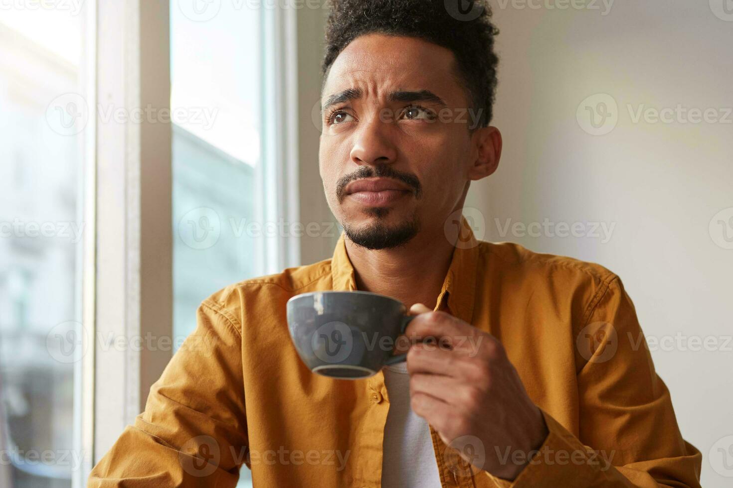 portrait de Jeune attrayant africain américain en pensant garçon, les boissons aromatique café de une gris tasse , en essayant à rappelles toi quelque chose et pensivement regards en haut. photo
