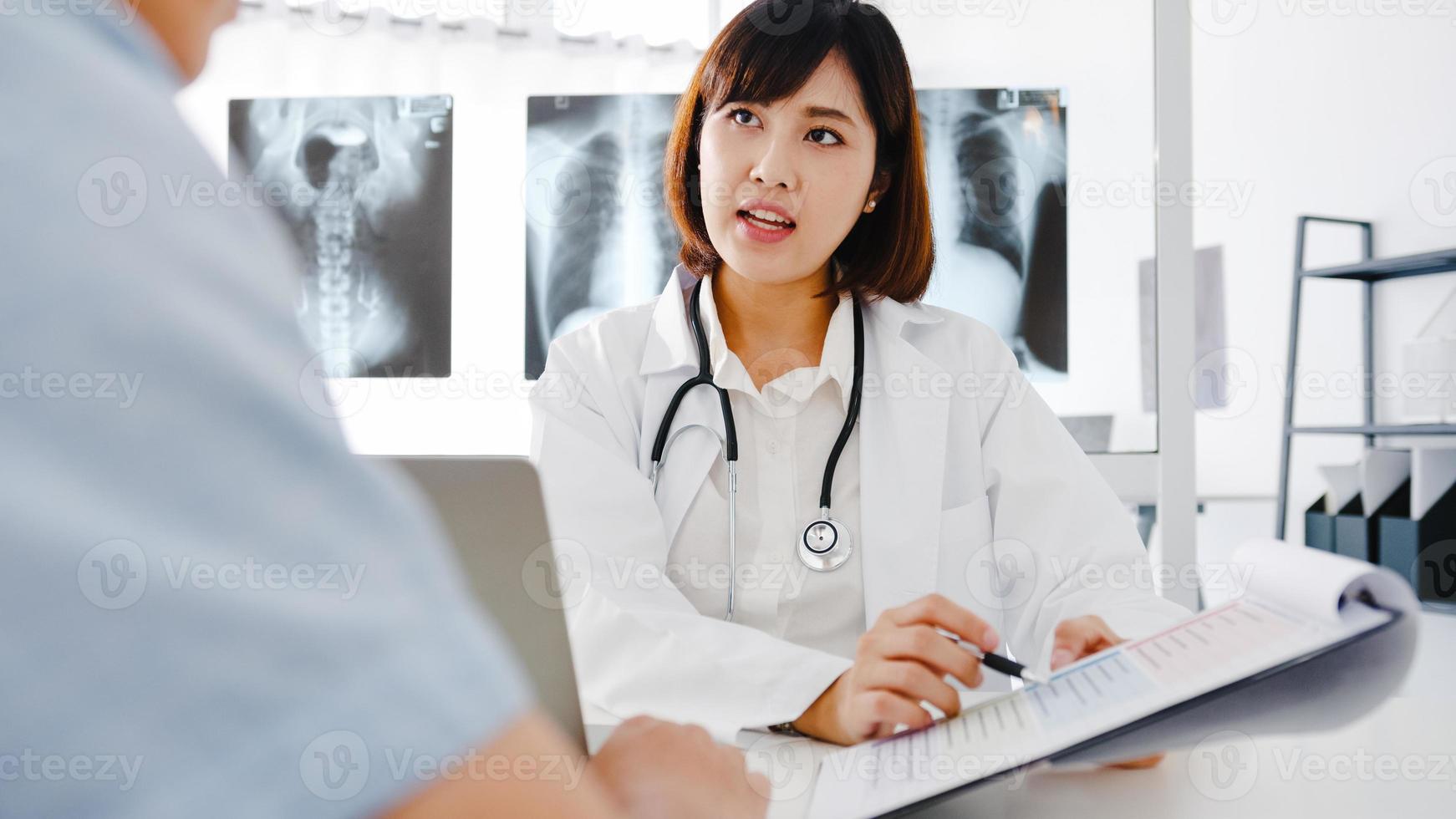 une jeune femme médecin asiatique en uniforme médical blanc utilisant un presse-papiers livre d'excellentes nouvelles discutent des résultats ou des symptômes avec un patient assis au bureau dans une clinique de santé ou un bureau d'hôpital. photo