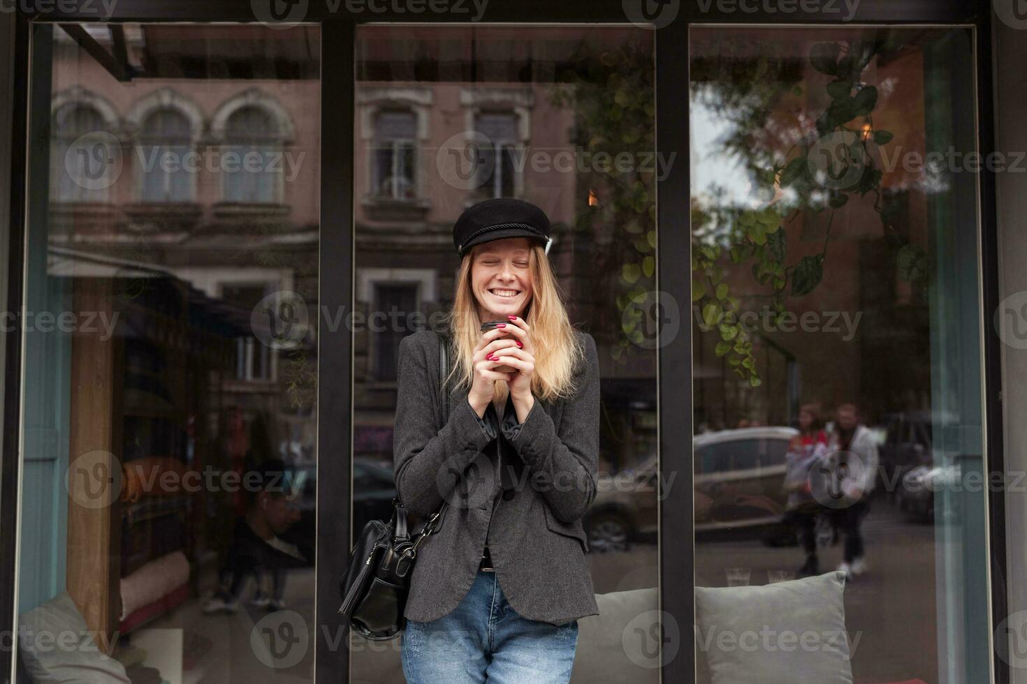 joyeux Jeune charmant blond longue aux cheveux Dame en portant tasse de café dans élevé mains et en riant Heureusement avec fermé yeux, posant Extérieur plus de gros fenêtre dans branché vêtements photo