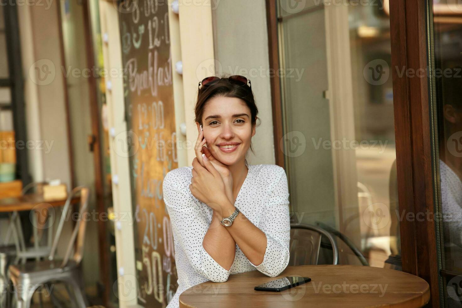 coup de Jeune magnifique aux yeux bruns brunette femme avec des lunettes de soleil sur sa tête en gardant mains sur table tandis que à la recherche gaiement à caméra, habillé dans blanc à pois porter photo