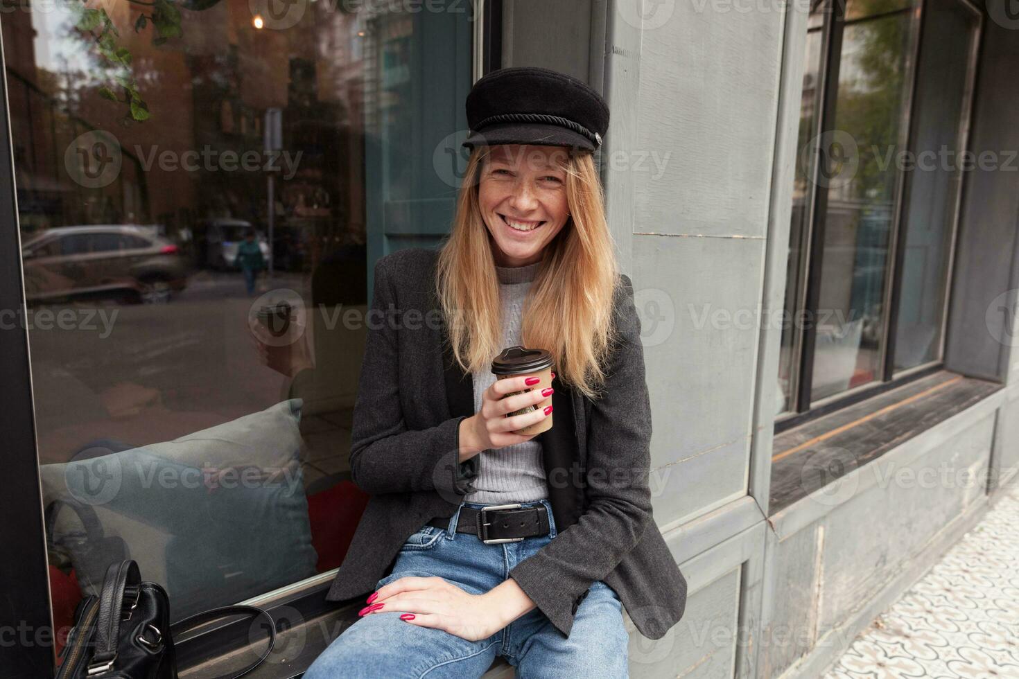 joyeux attrayant Jeune blond longue aux cheveux femme dans branché vêtements séance plus de gros café fenêtre avec tasse de café dans sa main, à la recherche à caméra avec large content sourire photo
