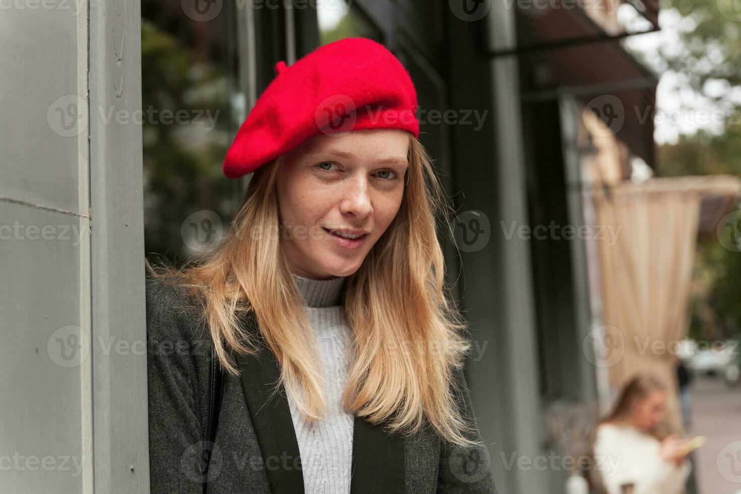 fermer de magnifique Jeune femme avec longue blond cheveux portant élégant vêtements tandis que posant plus de Urbain arrière-plan, à la recherche à caméra et souriant légèrement photo