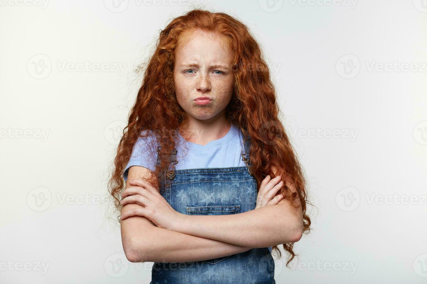 prétrait de malheureux taches de rousseur peu fille avec gingembre cheveux, des stands plus de blanc mur avec franchi bras, regards triste et offensé. photo