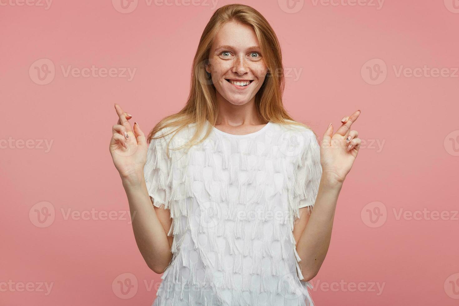 agité Jeune joyeux roux femelle avec Naturel maquillage élevage mains avec franchi les doigts et à la recherche avec enthousiasme à caméra avec vaste de bonne humeur sourire, permanent plus de rose Contexte photo