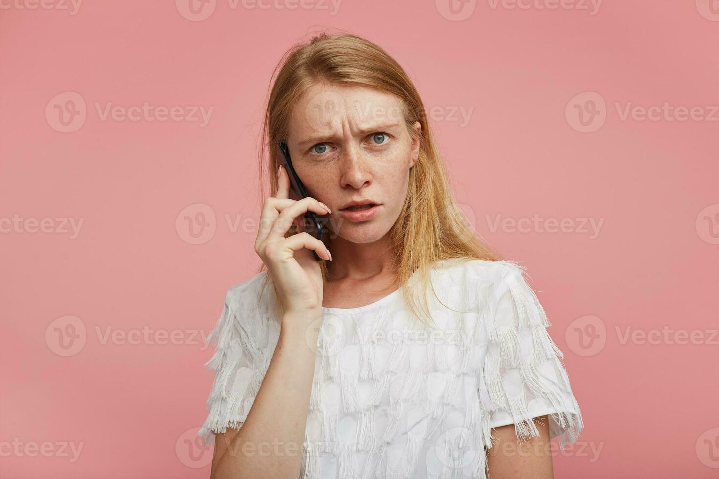 perplexe Jeune roux femme habillé dans blanc de fête T-shirt ayant désagréable téléphone conversation et à la recherche confusément à caméra, permanent contre rose Contexte photo