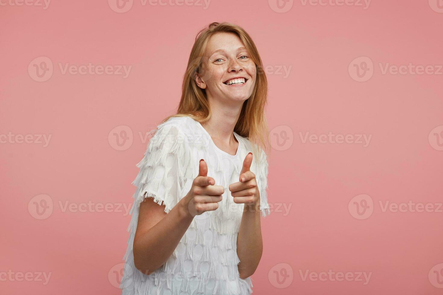 intérieur coup de Jeune content roux femme avec Naturel maquillage montrer du doigt à caméra avec élevé index et souriant gaiement tandis que posant plus de rose Contexte photo