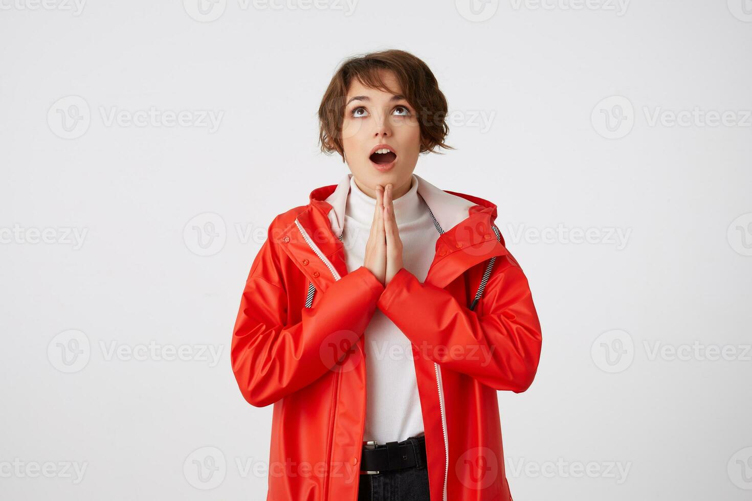 agréable positif Jeune court aux cheveux Dame dans blanc le golf et rouge pluie manteau, regards en haut avec large ouvert bouche à le copie espace, fait du prière geste et espère pour chance. permanent plus de blanc Contexte. photo