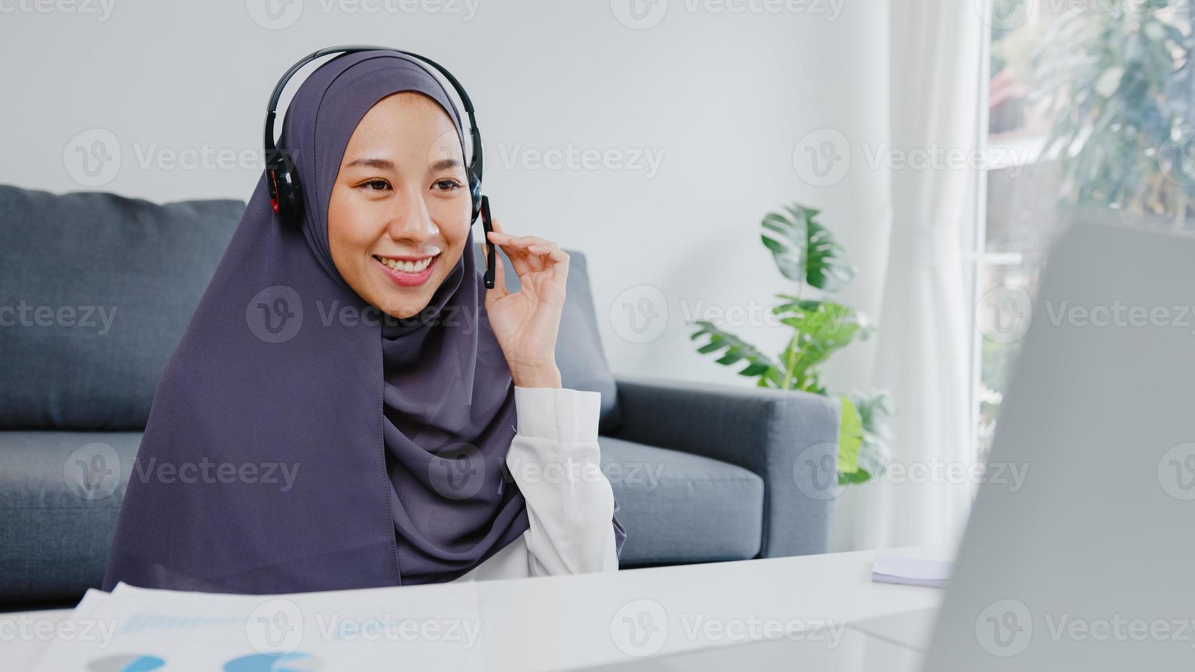 une dame musulmane d'asie porte un casque à l'aide d'un ordinateur portable parle à ses collègues du plan lors d'une conférence vidéo tout en travaillant à domicile dans le salon. distanciation sociale, quarantaine pour la prévention du virus corona. photo