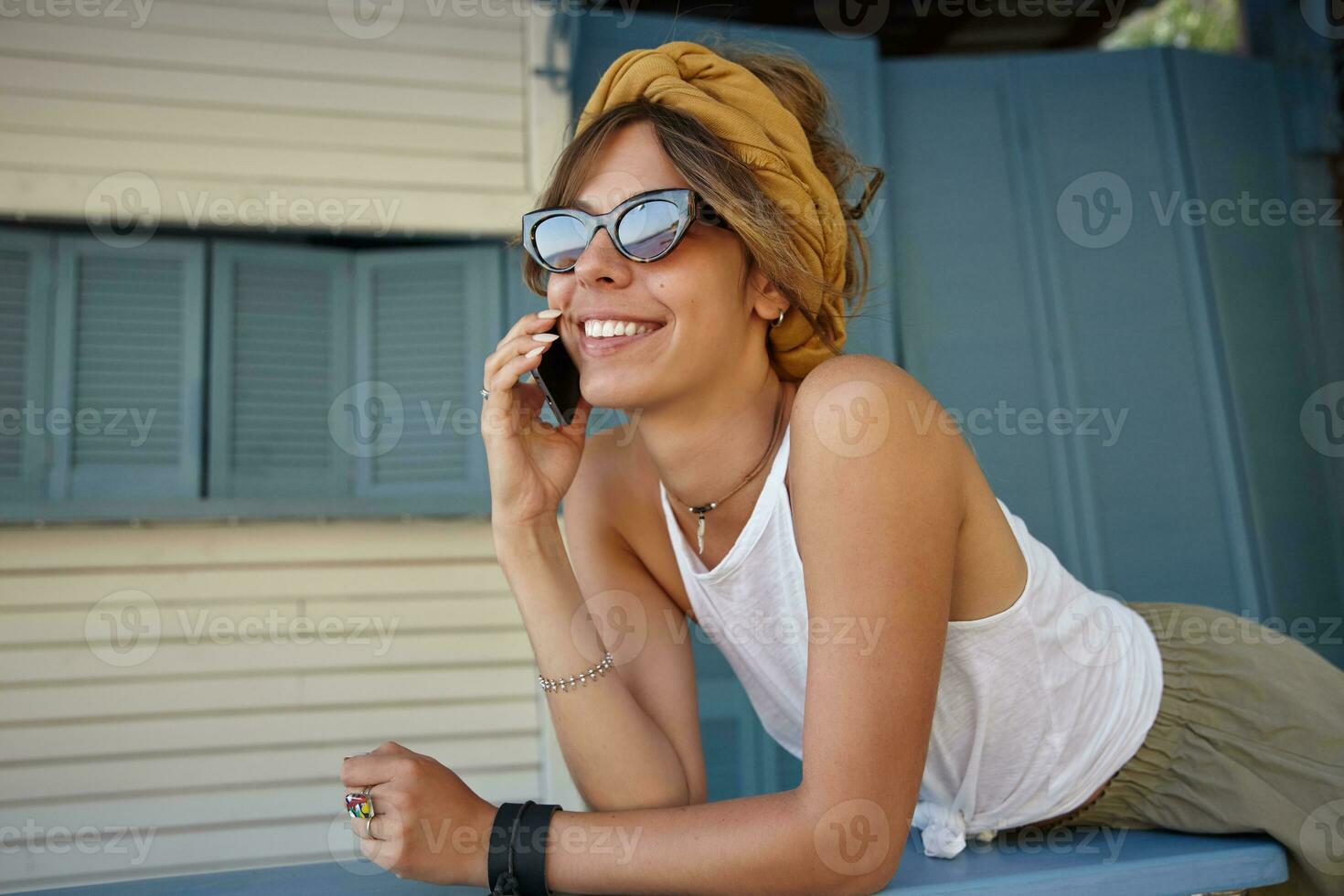 magnifique Jeune brunette Dame avec décontractée coiffure portant blanc Haut et olive jupe, posant plus de été terrasse sur chaud ensoleillé jour, fabrication appel avec sa mobile téléphone et souriant Cordialement photo