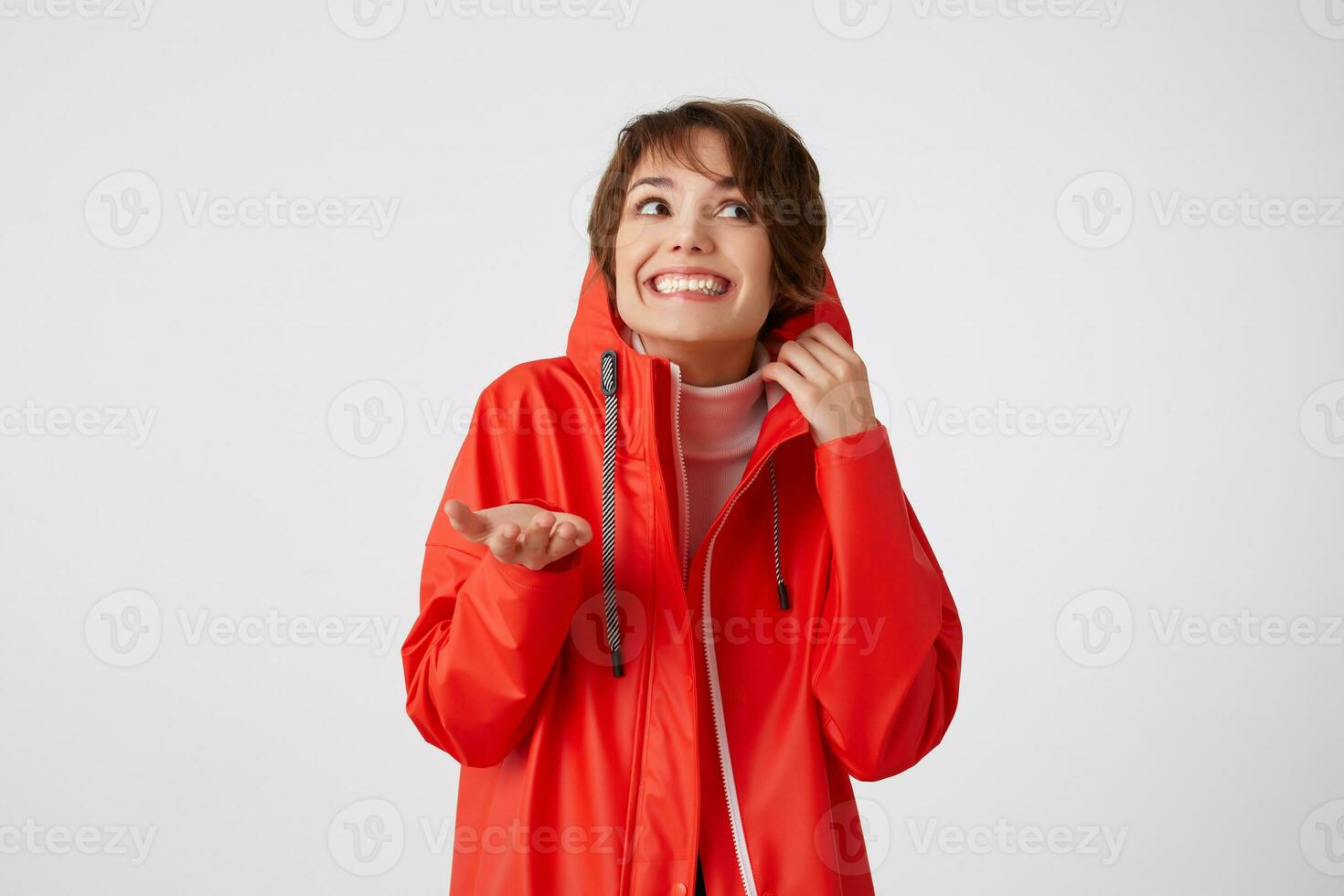 photo de beauté Jeune souriant court aux cheveux femme dans rouge pluie manteau, à la recherche en haut à le gauche, cache dans le capot, met le paume en dessous de le pluie.debout plus de blanc Contexte.