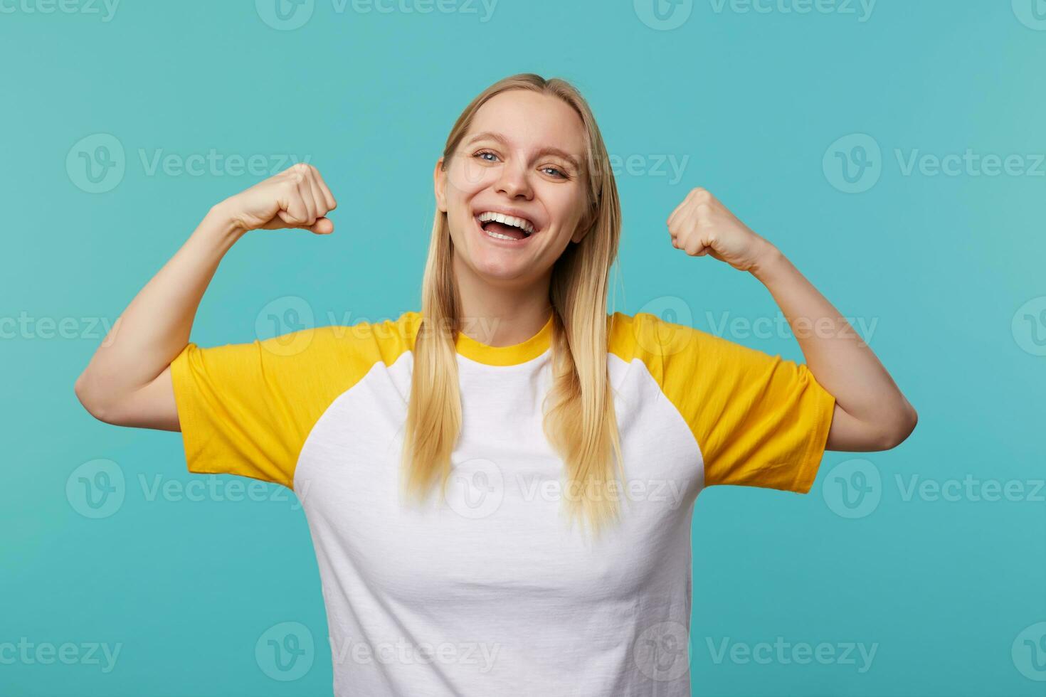 content Jeune aux yeux bleus longue aux cheveux blond femme à la recherche gaiement à caméra avec large sourire tandis que montrant fort biceps, permanent plus de bleu Contexte photo