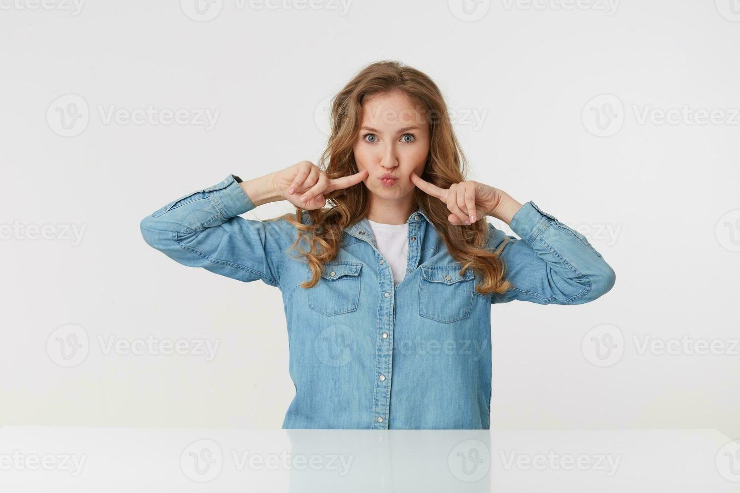 photo de Jeune mignonne femme avec longue blond ondulé cheveux dans denim chemise, touche le sien des joues et soufflant lèvres, regards drôle. isolé plus de blanc Contexte.