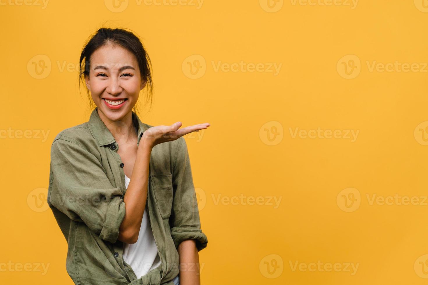 portrait d'une jeune femme asiatique souriante avec une expression joyeuse, montre quelque chose d'étonnant dans un espace vide dans un tissu décontracté et regardant la caméra isolée sur fond jaune. concept d'expression faciale. photo