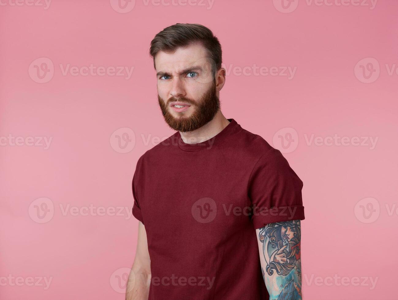 photo de Jeune Beau renfrogné malentendu rouge barbu homme dans Vide T-shirt, des stands plus de rose Contexte regards à le caméra avec dégoûter.