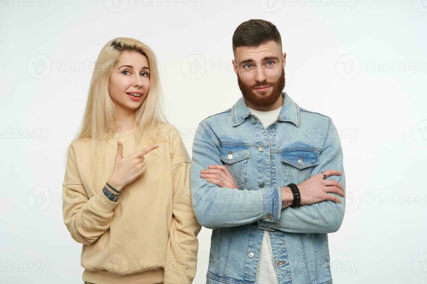 studio photo de Jeune jolie blond femelle dans beige sweat-shirt montrer du doigt positivement sur mignonne Jeune marron aux cheveux barbu homme avec plié mains, posant plus de blanc Contexte