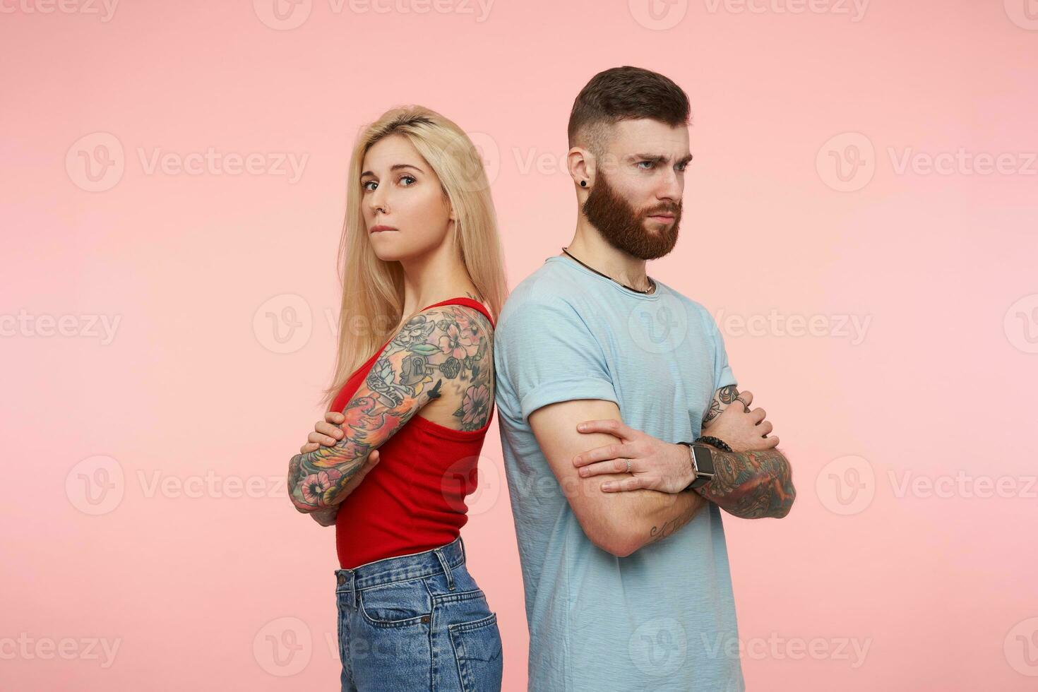 sérieux tatoué paire de jeune gens traversée mains sur coffres et renfrogné Malheureusement les sourcils, portant décontractée vêtements tandis que permanent plus de rose Contexte photo