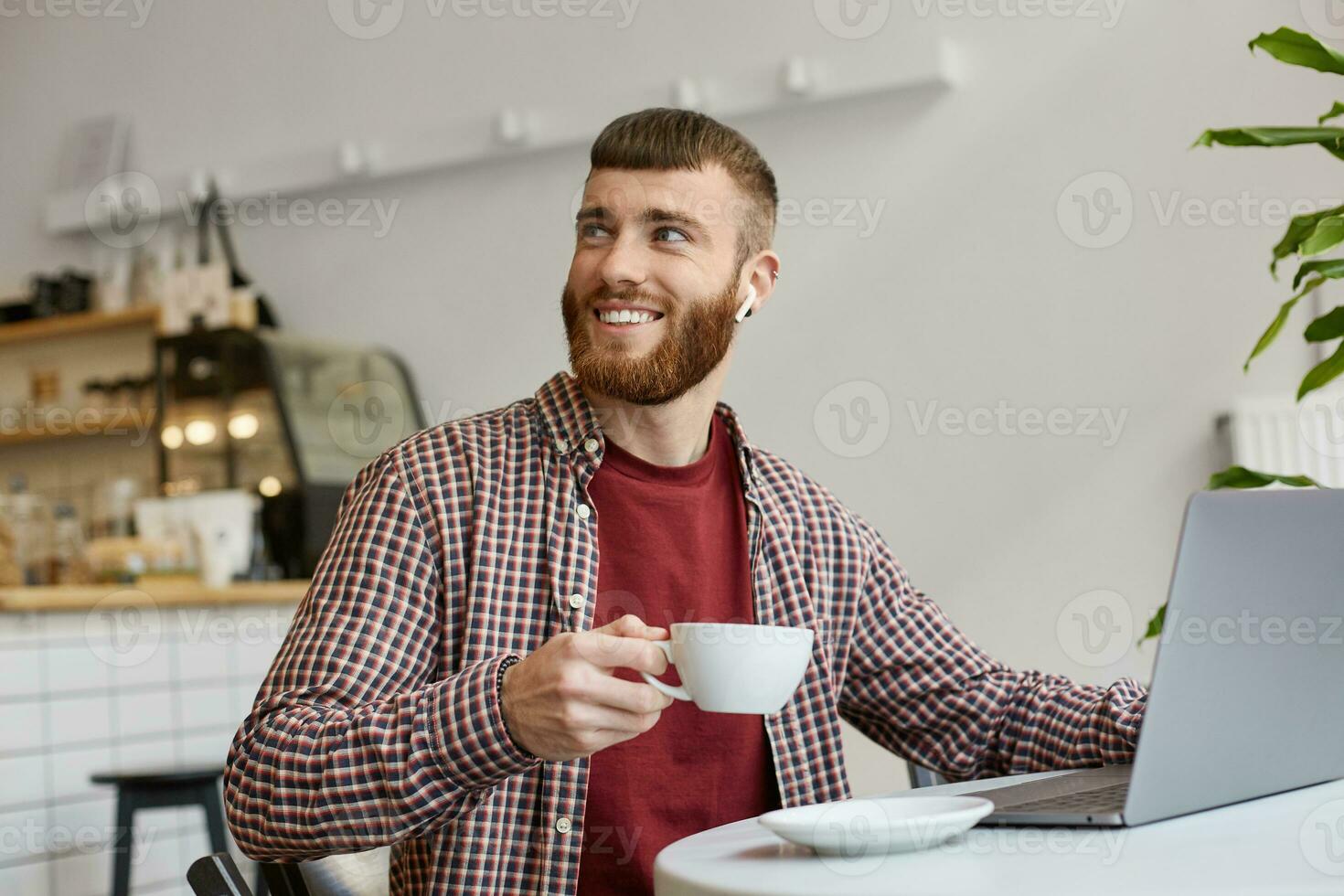 photo de content souriant attrayant gingembre barbu homme travail à une ordinateur portable, séance dans une café, en buvant café, portant dans de base vêtements, à la recherche à le droite, Merci barista pour une magnifique café.