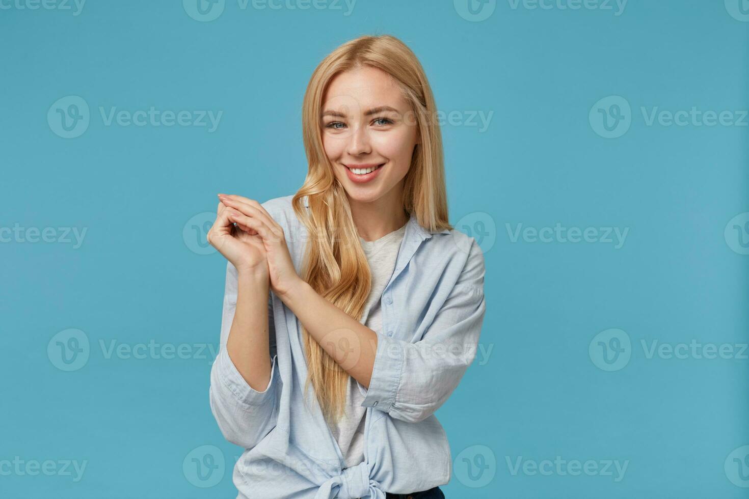 charmant Jeune blond longue aux cheveux femme élevage plié mains et souriant à caméra gaiement, permanent plus de bleu Contexte dans décontractée vêtements, démontrant sa blanc parfait les dents photo