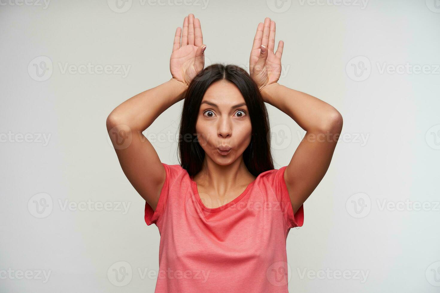 portrait de de bonne humeur Jeune marron aux cheveux femme avec décontractée coiffure en gardant élevé mains en dessous de sa tête et fabrication marrant visages tandis que permanent plus de blanc Contexte photo