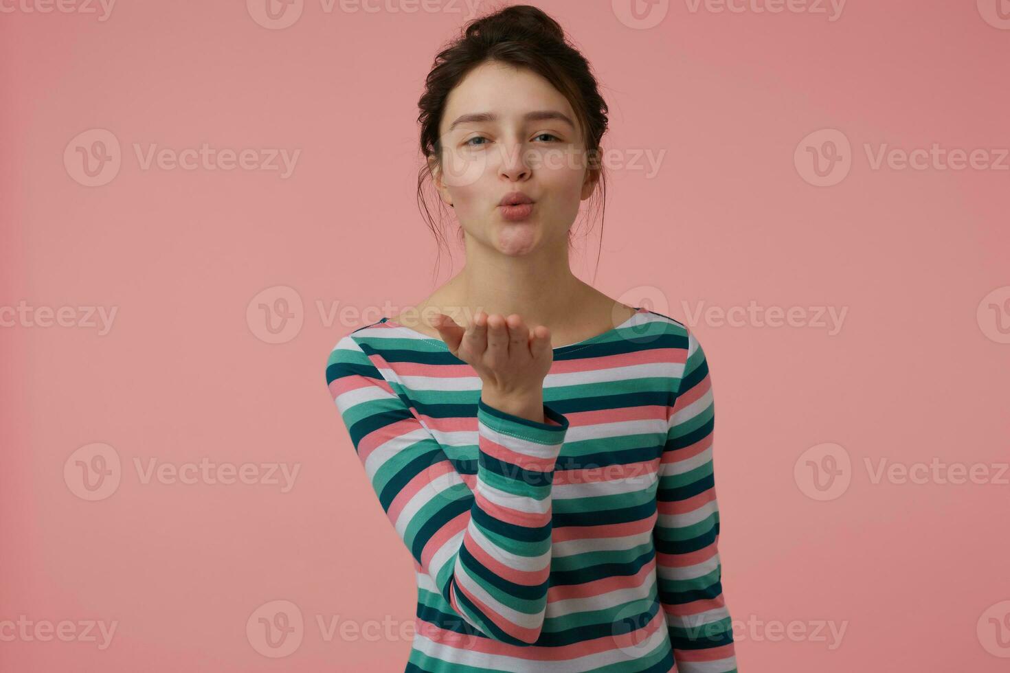 adolescent fille, content en regardant, charmant femme avec brunette cheveux et chignon. portant rayé chemisier et Envoi en cours une baiser. émotif concept. en train de regarder à le caméra isolé plus de pastel rose Contexte photo