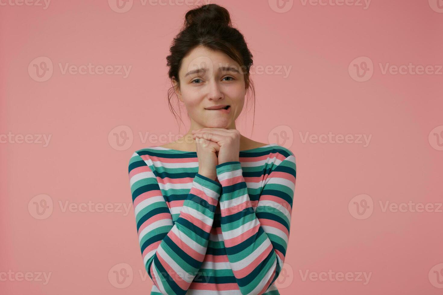 adolescent fille, content à la recherche femme avec brunette cheveux et chignon. portant rayé chemisier et mordant sa lèvre, demander. émotif concept. en train de regarder à le caméra isolé plus de pastel rose Contexte photo