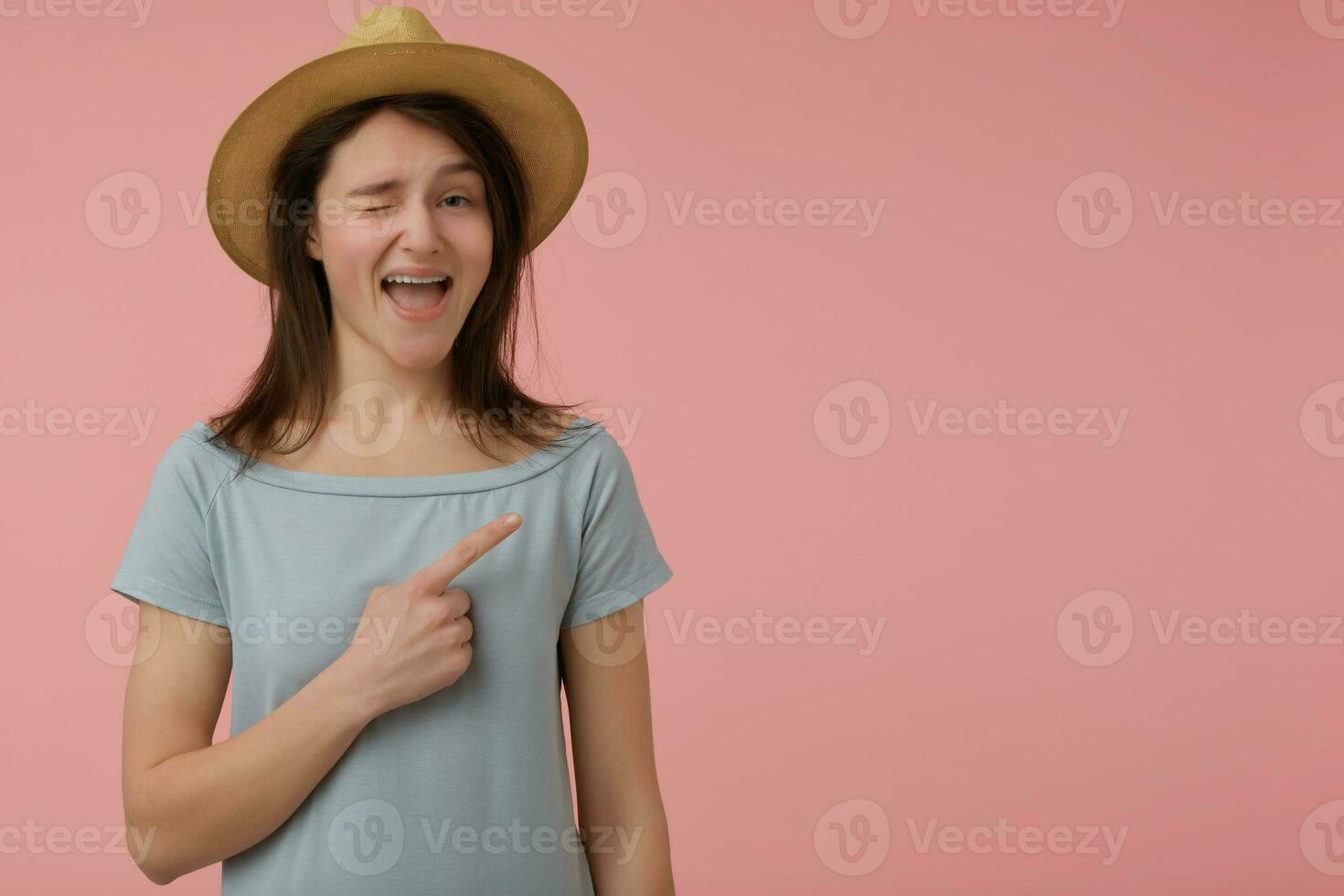 portrait de attractif, adulte fille avec longue brunette cheveux. portant bleuâtre T-shirt et chapeau. clin d'œil, souriant à le caméra et montrer du doigt à le droite à copie espace plus de pastel rose Contexte photo