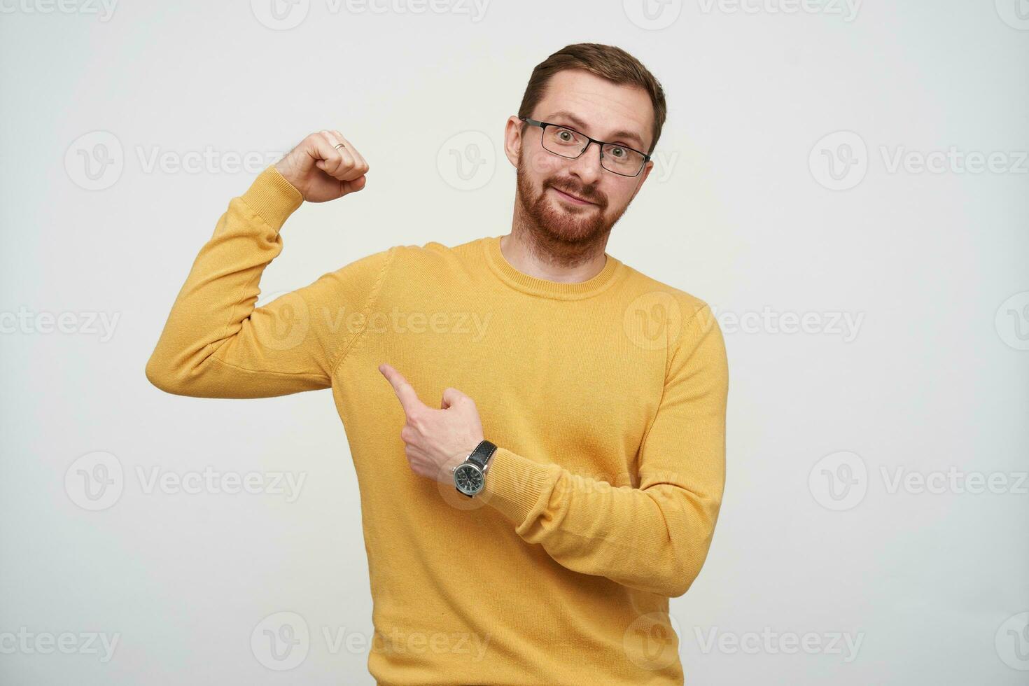 positif Jeune attrayant marron aux cheveux Masculin avec barbe portant lunettes et moutarde arrêtez-vous tandis que permanent plus de blanc arrière-plan, montrant sur le sien biceps avec indice doigt et souriant légèrement à caméra photo