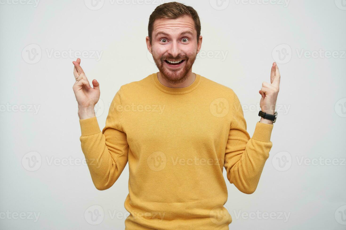 fou de joie attrayant Jeune barbu homme avec marron court cheveux à la recherche à caméra avec large yeux et bouche ouvert et traversée le sien les doigts pour bien chance, permanent contre blanc Contexte photo